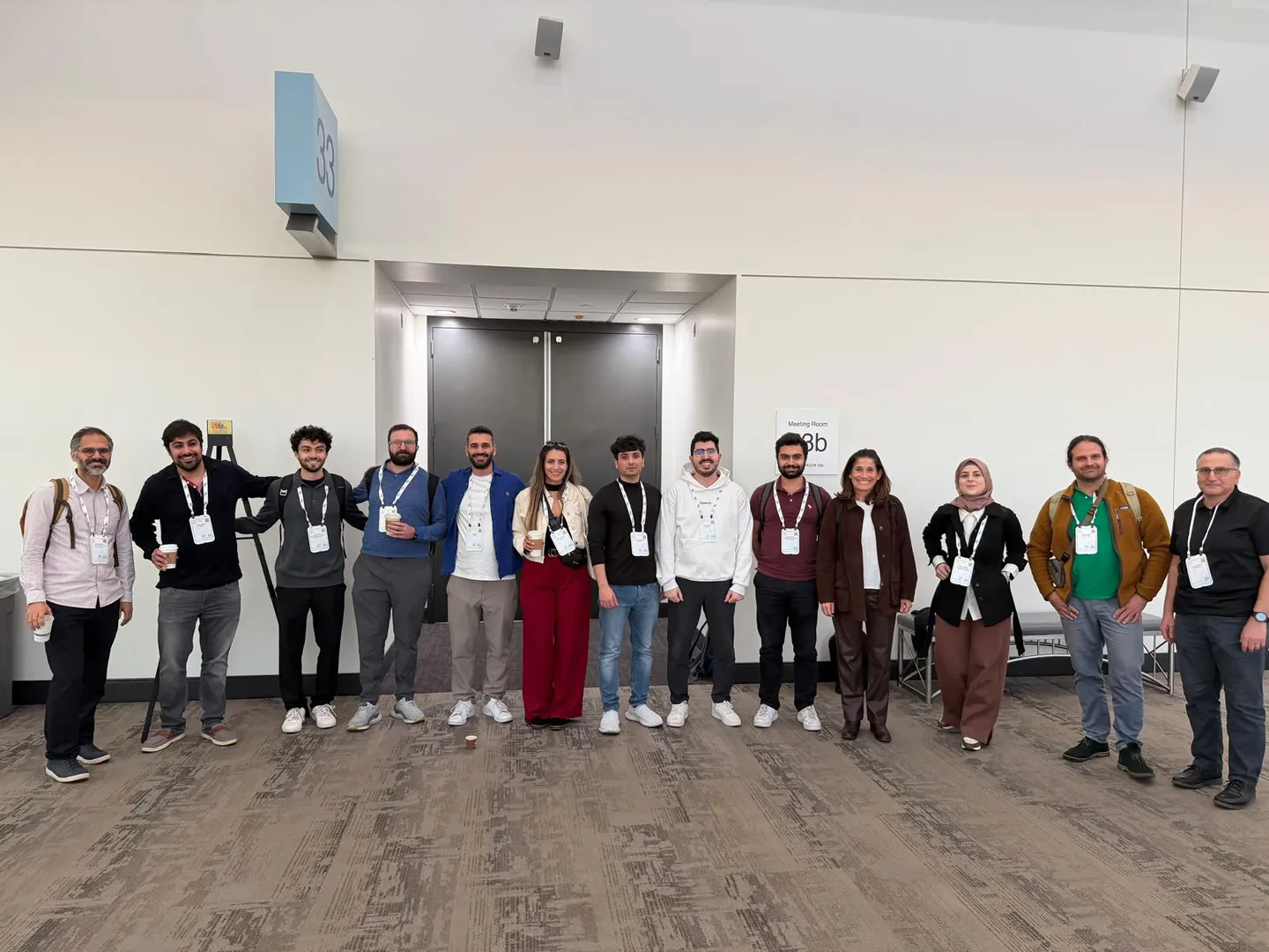 Thirteen conference attendees pose in a line outside meeting room 3B at NeurIPS 2025